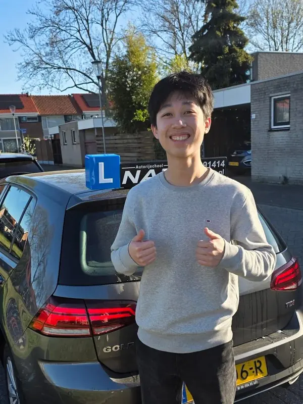 Geslaagde leerling steekt beide duimen op bij de lesauto van NAZ